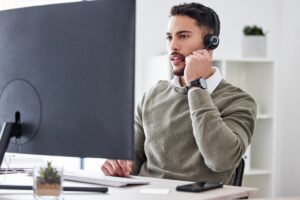 Man sitting on desk at office space while holding headset and working with computer monitor.