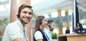 Male telemarketer smiling while working on office space with other blurred telemarketers on the background.