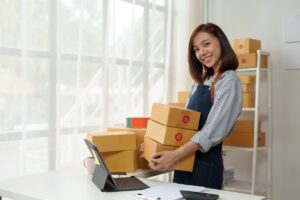 Woman wearing an apron is carrying cardboard boxes in storage room. Laptop on desk.