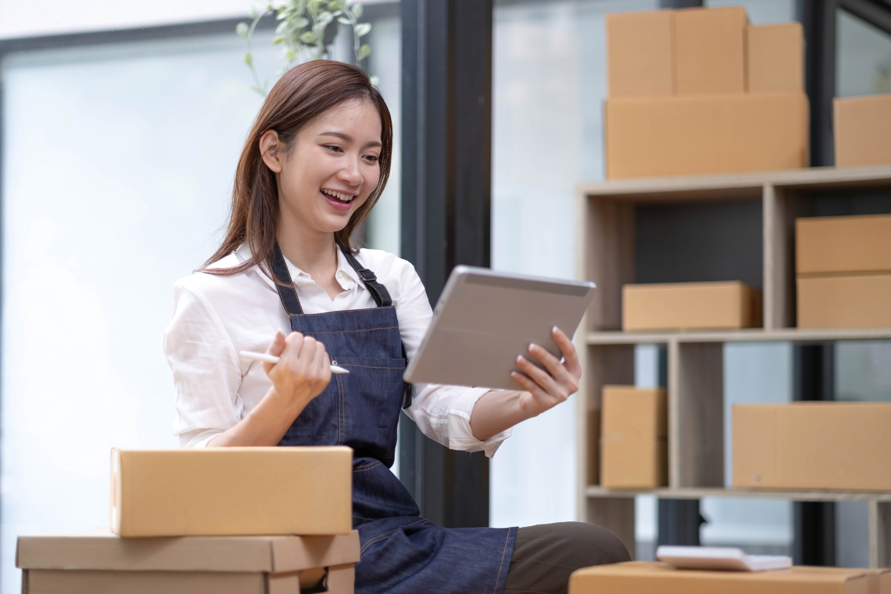 A smiling woman in an apron holding a tablet while working in a small business setup, surrounded by cardboard boxes.