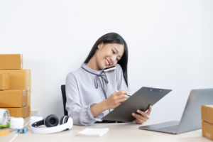 A cheerful young woman multitasking by talking on the phone while writing on a clipboard, surrounded by cardboard boxes and a laptop, representing a busy e-commerce or small business environment.