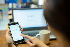 Woman holding phone while searching on google, blurred laptop on the background.