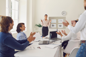 Businesswoman giving a presentation to colleagues in a meeting room while the team applauds.