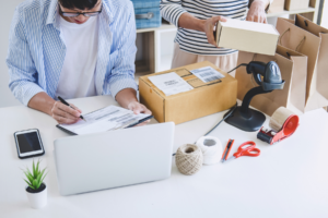 Two individuals organizing packages at a desk with shipping supplies and a laptop.