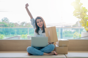 Woman sitting with a laptop, raising one hand in excitement, holding a package in the other, surrounded by boxes on a bright balcony.