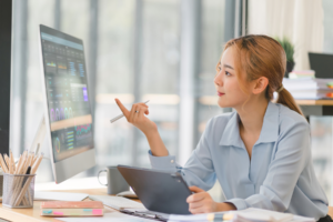 Woman sitting in office space while she points at computer screen as she hold tablet.