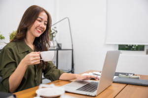 Smiling woman in a green shirt holding a cup of coffee while using a laptop at a wooden desk in a bright room.