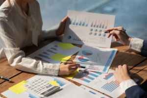 A close-up of two people discussing colorful charts and graphs during a business meeting, with a calculator and sticky notes on the wooden table.