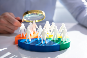 A person holding a magnifying glass over a colorful pie chart with small figurines representing people, symbolizing market segmentation or customer analysis