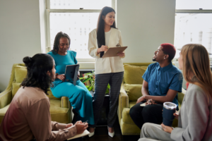 Five individuals seated on green couches in a meeting room, with one standing and presenting using a clipboard, while others are engaged in discussion.