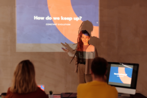 A woman giving a presentation with a projected slide behind her, titled "How do we keep up? Constant Evolution" while the audience listens attentively.