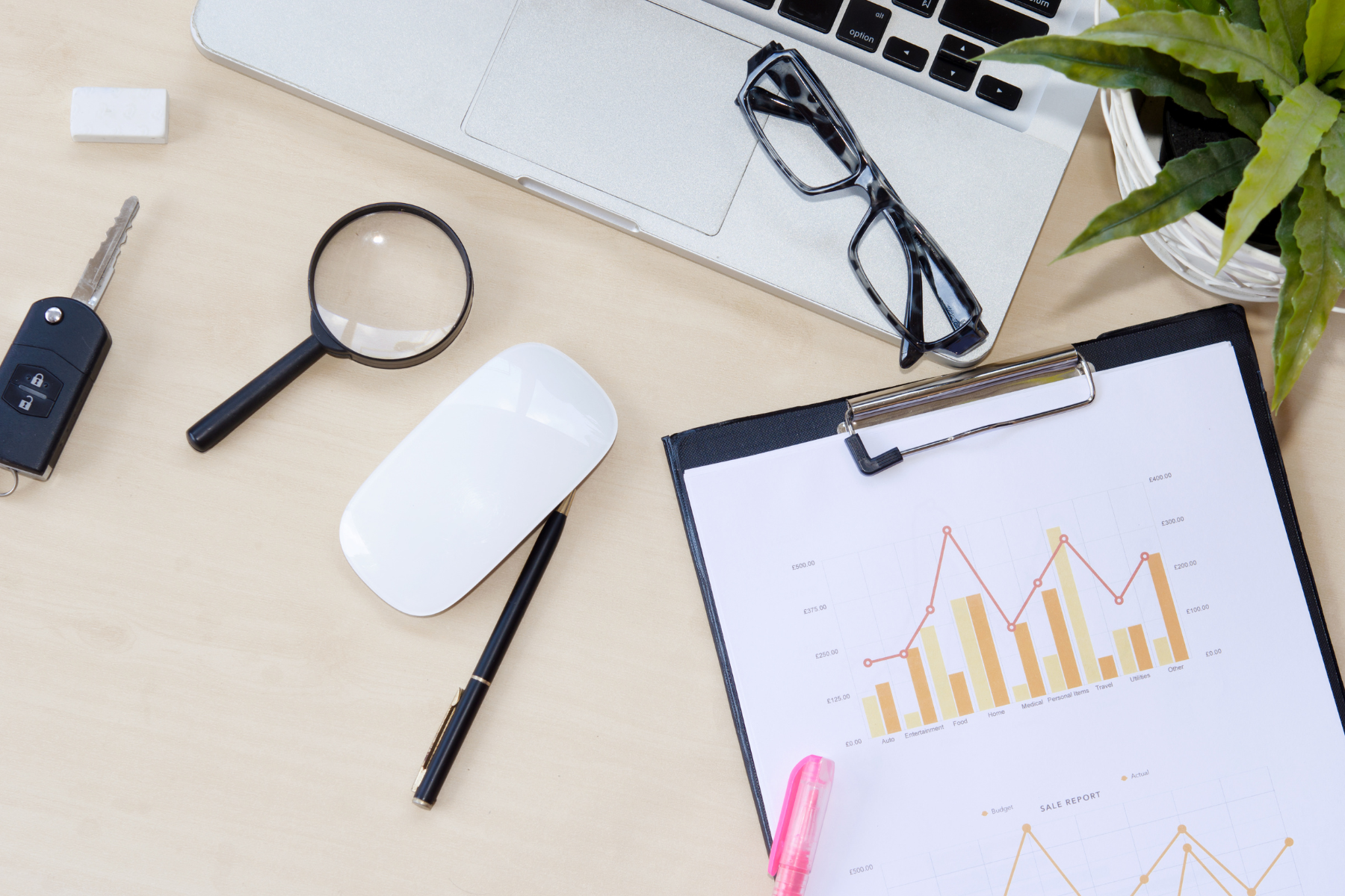 A desk setup with charts and graphs on paper, a magnifying glass, a mouse, glasses, keys, and a laptop, suggesting financial or data analysis.