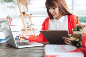 A woman in a red cardigan working on a laptop with a clipboard in hand, surrounded by small packages, indicating an e-commerce business or order management.