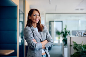 A smiling woman in a gray blazer standing with arms crossed in a modern office, representing confidence and career success.