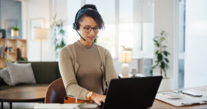 A friendly woman wearing a headset, working on her laptop in a home office, representing remote customer service or sales support.