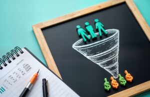 A blackboard with a hand-drawn sales funnel illustration, featuring human figures entering the funnel and dollar signs at the bottom, symbolizing lead generation and conversion in marketing. A notepad and pens are placed beside it, representing strategic planning and business growth.