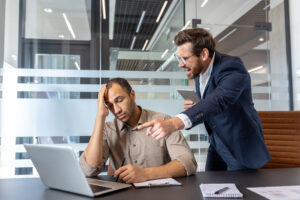 A tense moment in an office where a man in a blue suit is visibly angry, pointing towards his coworker, who appears stressed and is holding his head in his hand. The interaction seems confrontational, and the environment looks modern with glass walls and office furniture.