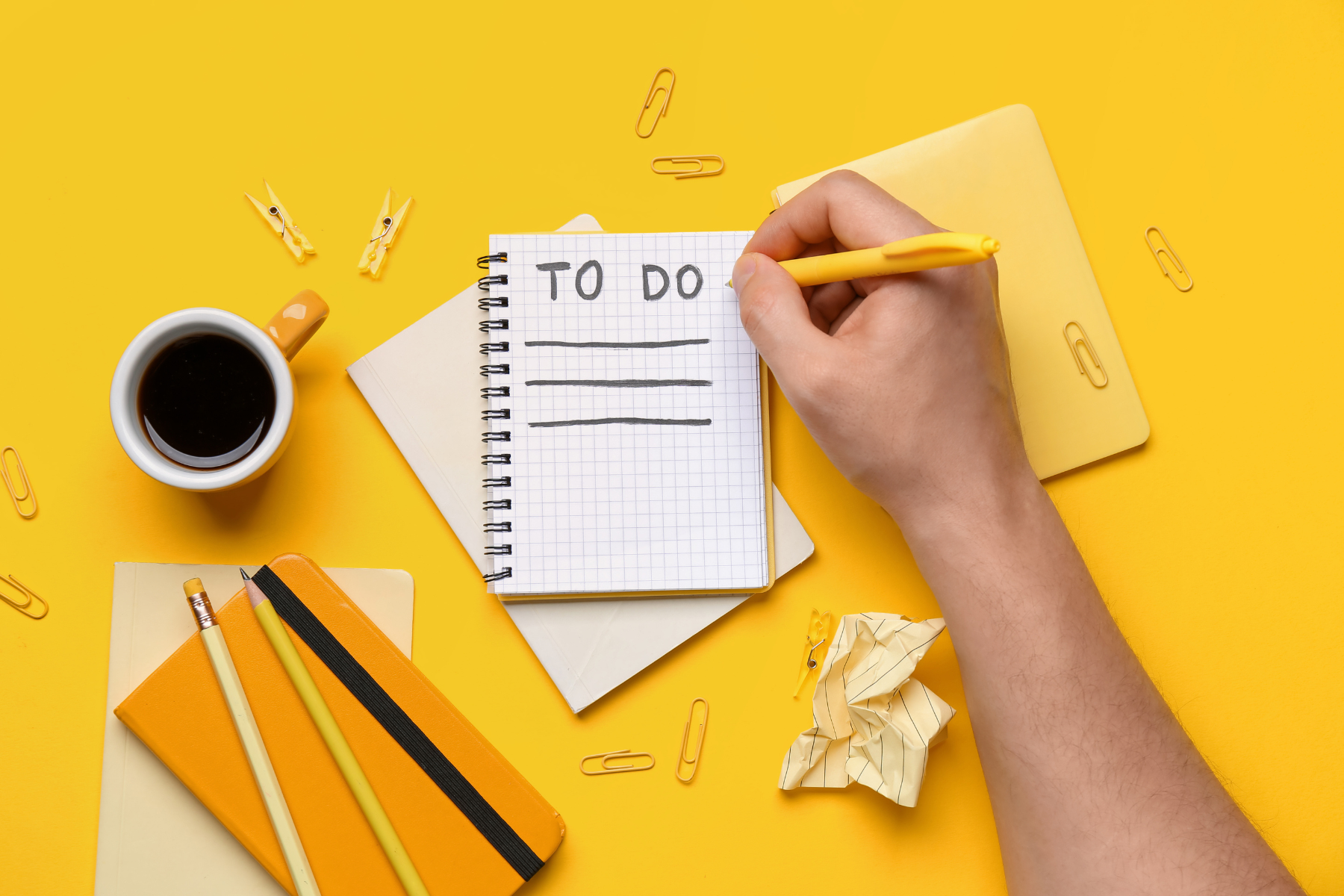 A hand holding a yellow pen and writing in a notebook on a bright yellow background. The page of the notebook has a "TO DO" header with three blank lines underneath it. Other stationery items like paper clips and notepads surround the scene.
