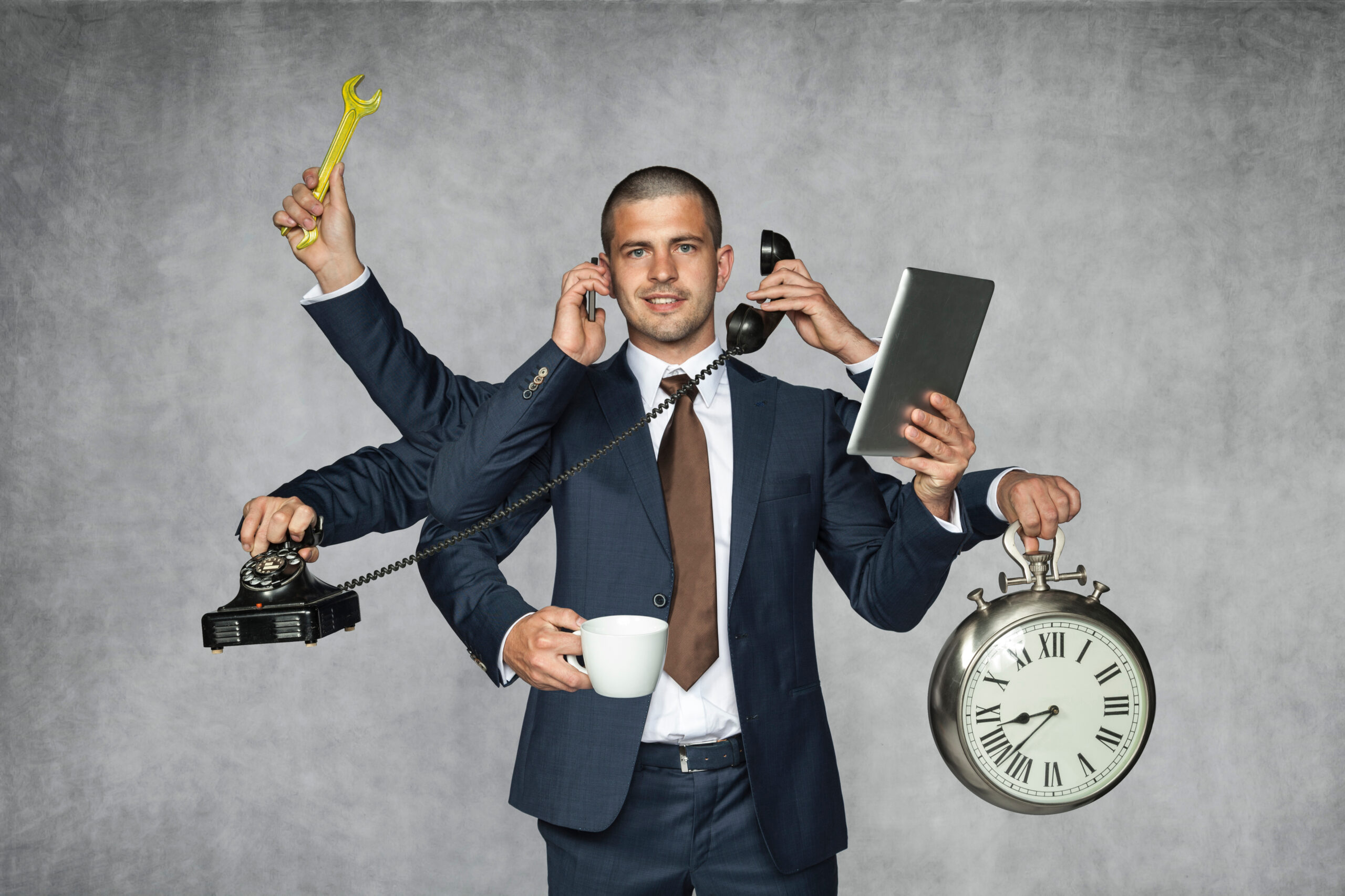 A man in a business suit is multitasking with four different hands engaged in various activities: one holding a wrench, another on the phone, a third holding a cup, and the last holding a tablet and an old-fashioned alarm clock. The image humorously illustrates a high-pressure work environment.