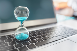 A close-up shot of an hourglass with blue sand positioned on top of a laptop keyboard. The sand in the hourglass is gradually running out.