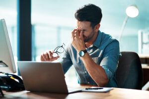 A man in a blue shirt is sitting at a desk, looking frustrated and rubbing his eyes while holding his glasses. He seems to be experiencing stress related to his work as he is surrounded by a computer and paperwork in a well-lit office.