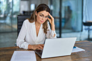 A woman wearing glasses and a white blouse is focused on her laptop in a modern office environment. She appears stressed as she rests her head on her hand, possibly overwhelmed by the task at hand.