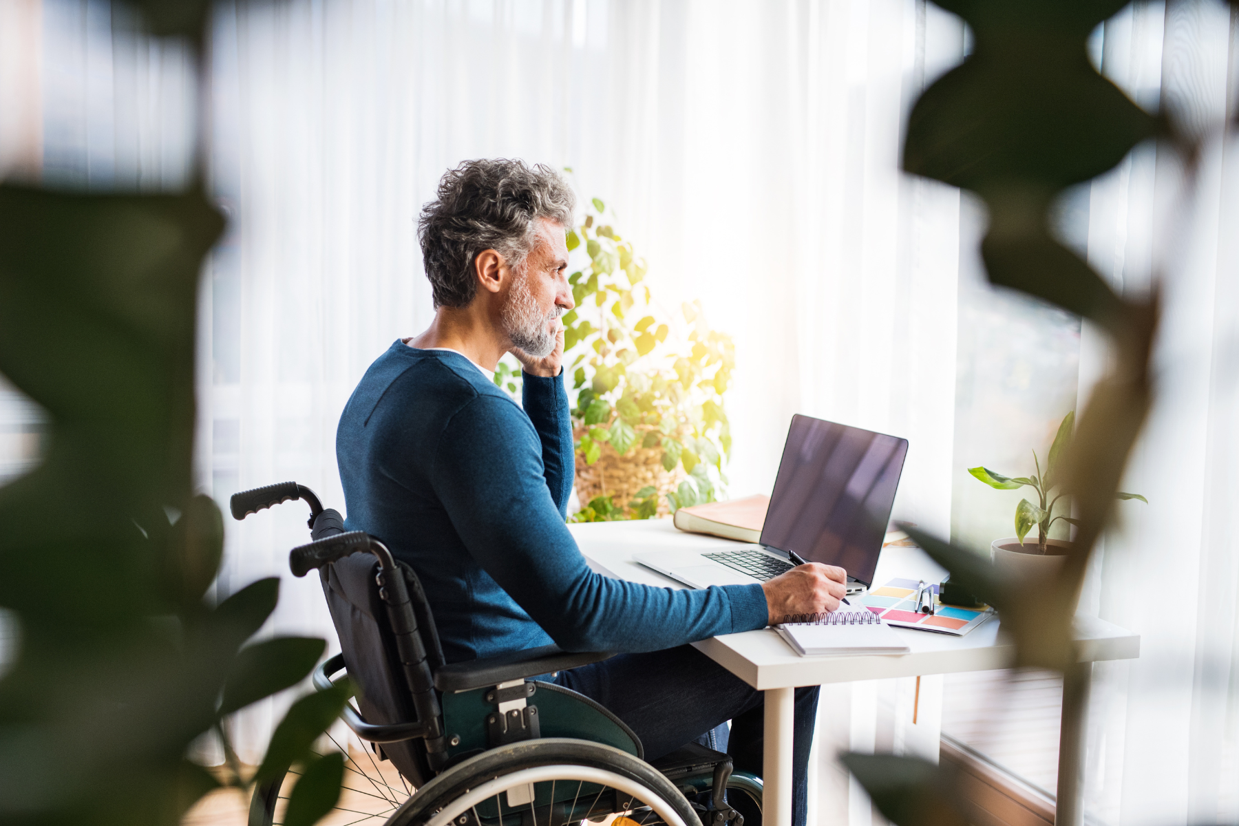 An older man with a beard is sitting in a wheelchair at a desk, working on a laptop. He appears to be in a thoughtful pose, resting his chin on his hand while looking at the screen. The desk is surrounded by plants, and natural light is streaming in through the window, creating a serene working environment.