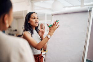 A woman gestures with a pen at a flipchart while speaking to someone out of frame, with some writing visible on the board.