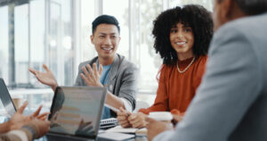 A group of professionals sit at a table in a meeting room, actively engaging in conversation with laptops and notebooks visible.