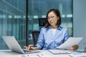 A woman in glasses sits at a desk, using a laptop and reviewing a document in a modern office setting.
