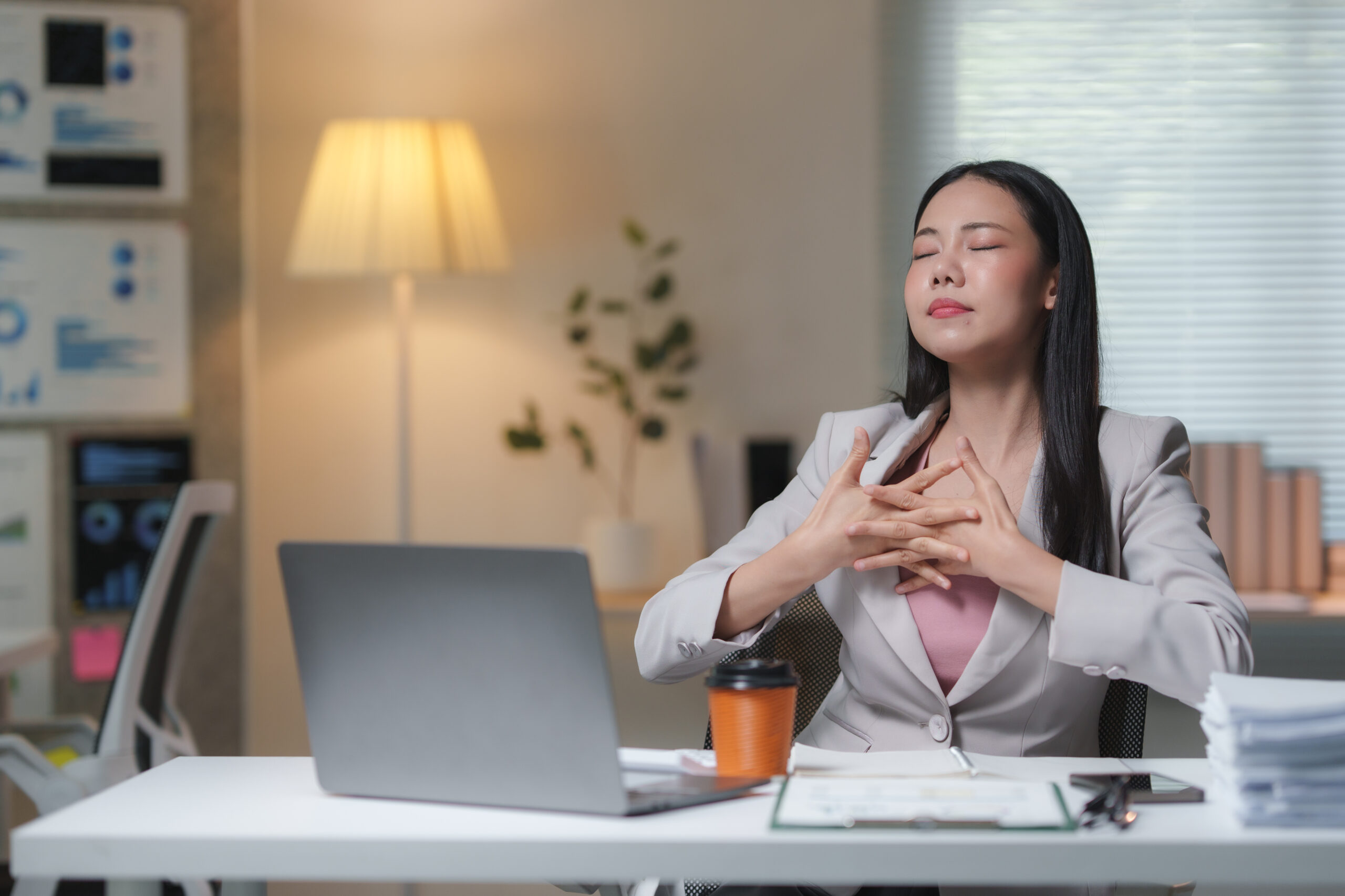 A woman in a suit sits at her desk with closed eyes and interlaced fingers, appearing relaxed, with a coffee cup and laptop in front of her.