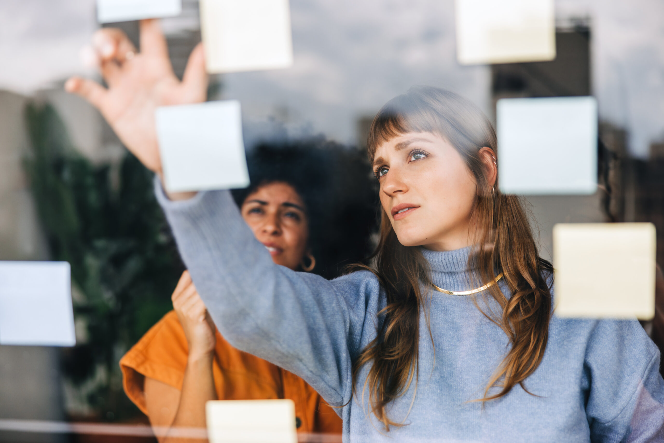 Two women stand behind a glass wall covered in sticky notes, with one reaching to place or adjust a note.
