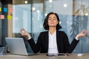 A woman in a black blazer sits cross-legged at a desk, eyes closed and hands in a meditative pose, with a laptop and phone nearby.