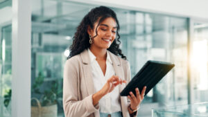 A smiling woman in business attire stands in an office, holding and tapping on a digital tablet.
