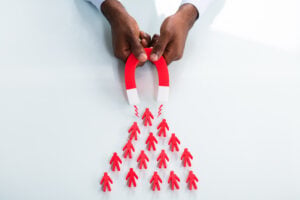 Close-up Of A Human Hand Attracting Red Human Figures With Horseshoe Magnet On White Background