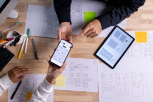 Designers collaborating on mobile app wireframes, reviewing interface mockups on a phone and tablet.