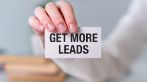 A close-up of a hand holding a small card that reads “GET MORE LEADS” in bold black text. The soft, blurred background suggests a direct call-to-action or promotional message.