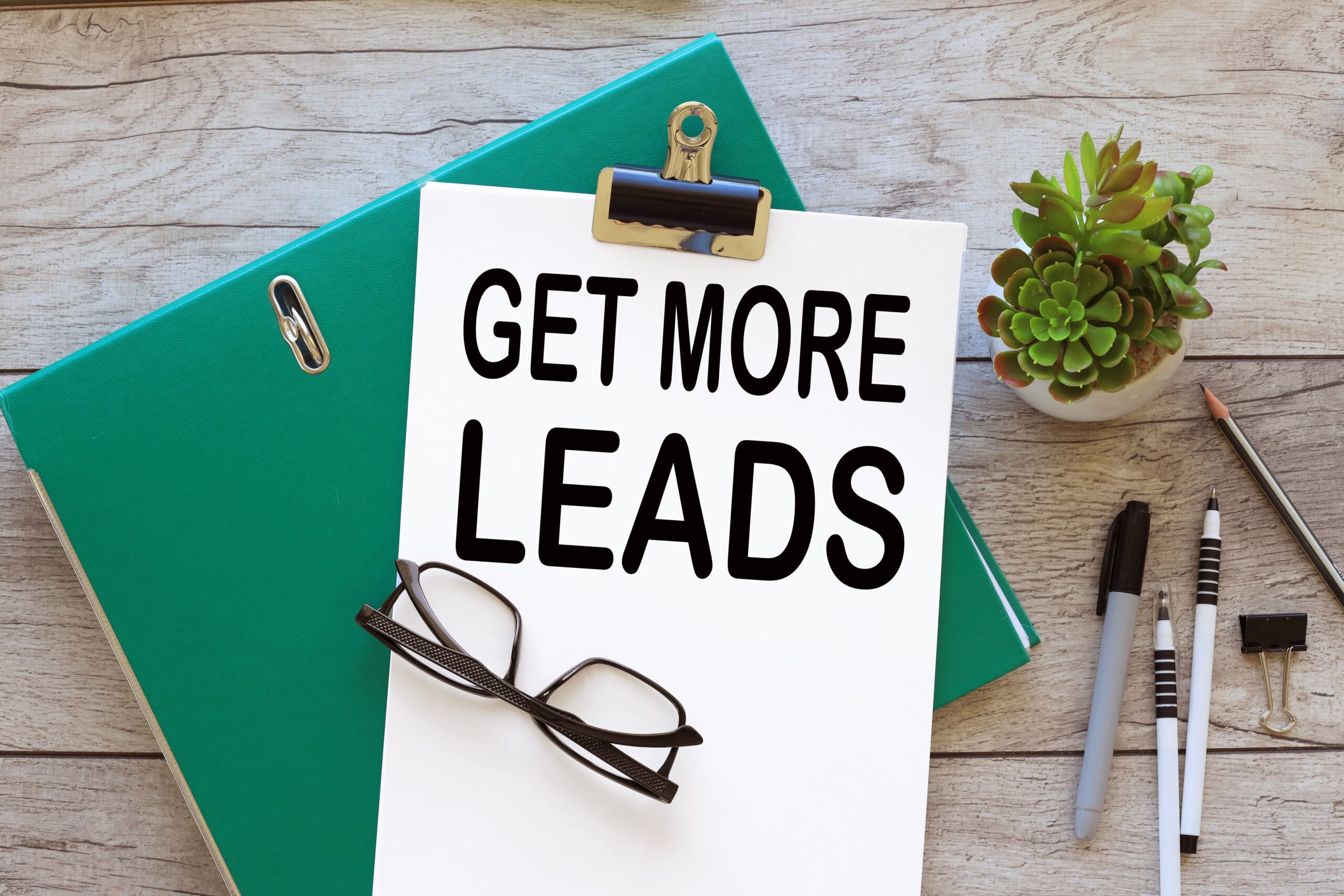 A flat-lay workspace image featuring a clipboard with a white sheet reading “GET MORE LEADS” in bold black text. It sits on a wooden desk alongside a green folder, eyeglasses, pens, a binder clip, and a small potted succulent—suggesting marketing planning or business growth.