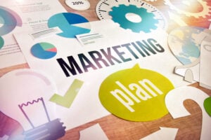 A colorful tabletop layout of marketing visuals with charts, icons, gears, and speech bubbles. The words “MARKETING plan” stand out, indicating structured planning, analytics, and strategy development.