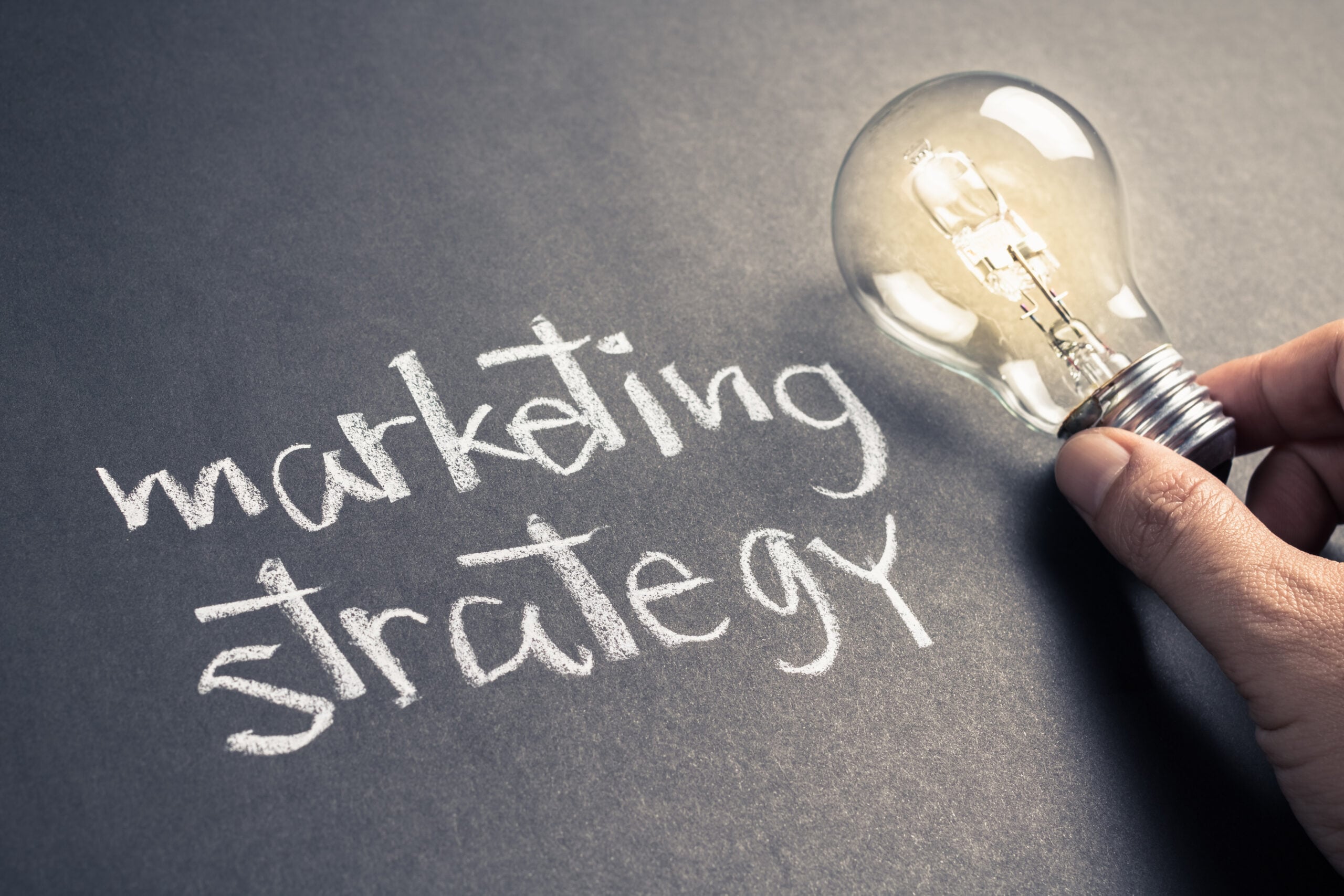 A close-up of a hand holding a light bulb over a chalkboard-style background with the words “marketing strategy” written in chalk. It symbolizes ideas, planning, and creative thinking in marketing.