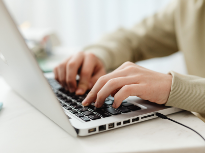 Close-Up of Hands Typing on a Laptop A close-up view of two hands typing on a silver laptop keyboard. The person wears a light green sweater, and the desk surface and blurred background are visible around them.