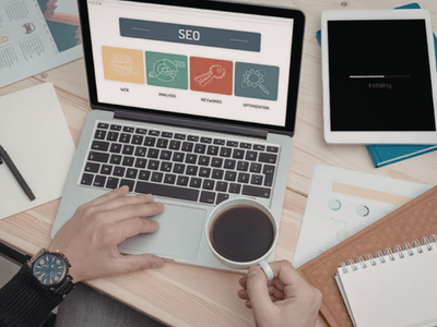 An overhead view of a desk where a person works on a laptop displaying an SEO dashboard, with a cup of coffee, a tablet and notebooks around it.