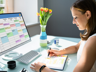 A woman at a desk writes in a paper planner while a weekly calendar is displayed on the monitor beside her.