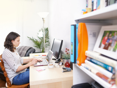 A woman sits at a home office desk typing a blog post on a laptop, with a monitor and a shelf of books beside her.